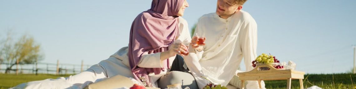 Muslim woman and man enjoying a sunny day picnic with snacks and drinks in a field.