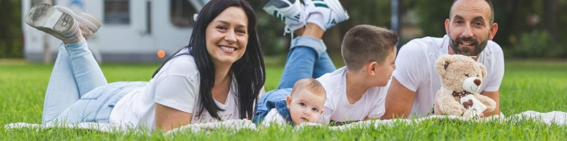 Happy family lying on grass with RV in the background, enjoying a sunny outdoor picnic.
